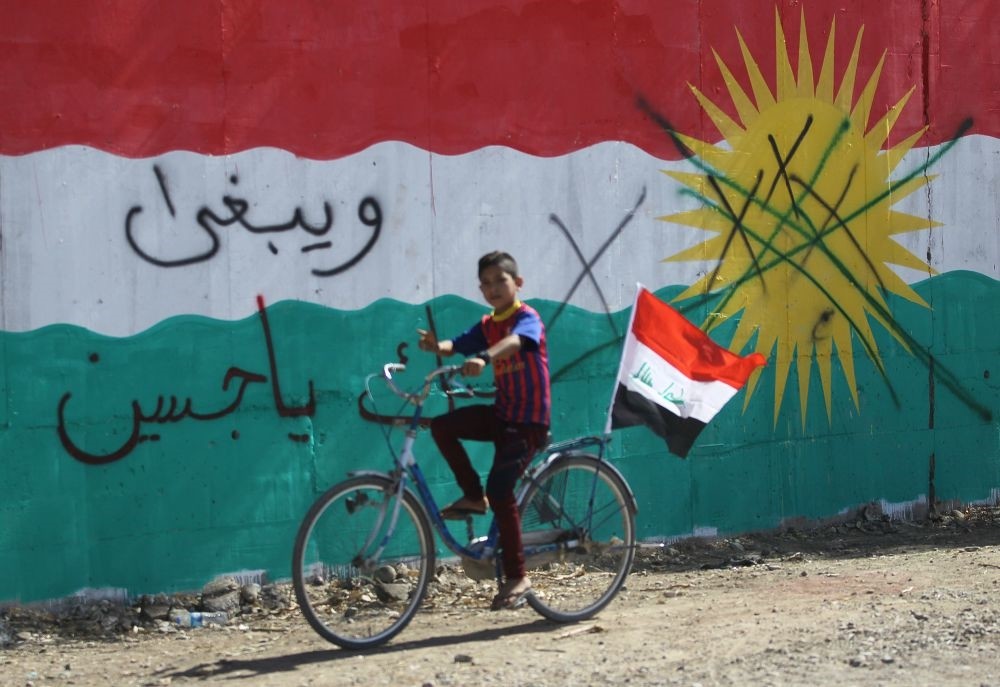 A boy rides a bicycle flying the Iraqi national flag past a wall mural depicting the KRG flag in the area of Dibs, west of Kirkuk on Oct. 17, 2017.