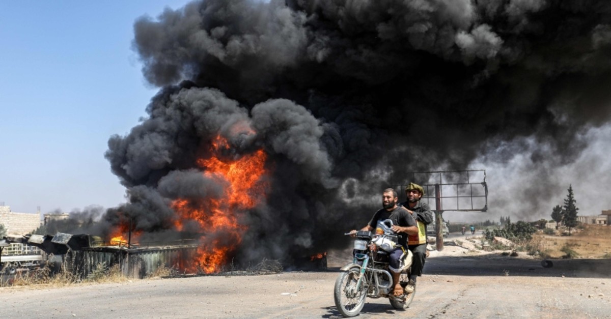 Men ride a motorcycle past a plume of smoke from a blaze at a vehicle gathering point for civilians fleeing from the south of Idlib province, which was hit by reported government forces' bombardment (AFP Photo)