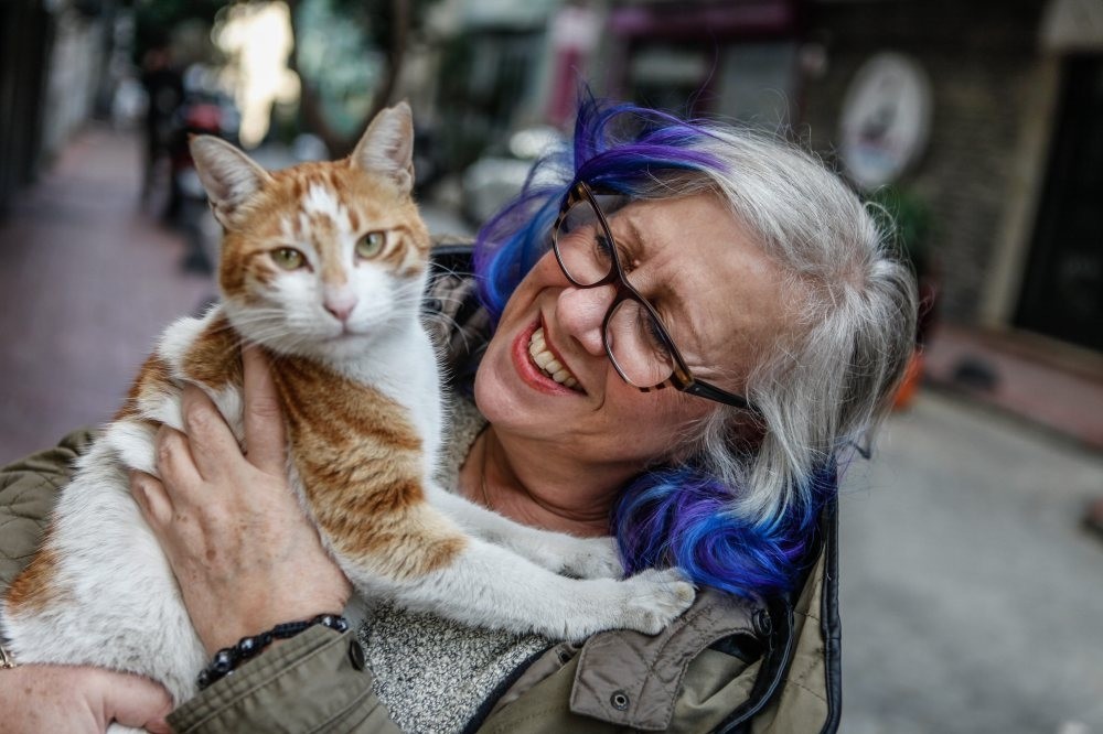 A woman with blue-tinted hair and glasses smiling while holding a ginger-and-white street cat on a quiet street in Cihangir.