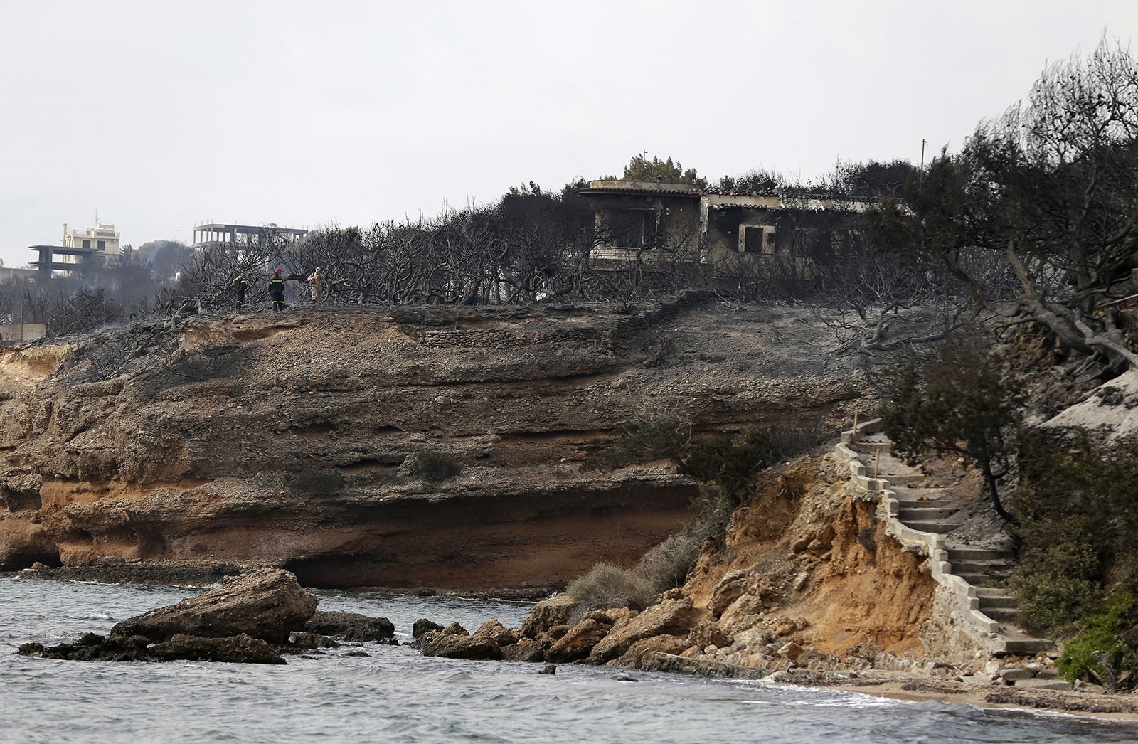 Firefighters stand on a cliff top where burned trees hug the coastline in Mati east of Athens, Tuesday, July 24, 2018.