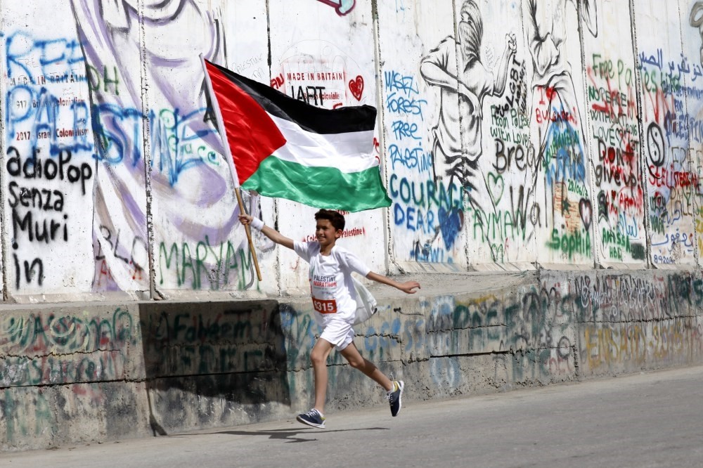 A child runs holding the Palestinian flag as he passes graffiti on the controversial Israeli separation barrier during the Palestine Marathon, Bethlehem, March, 2018.