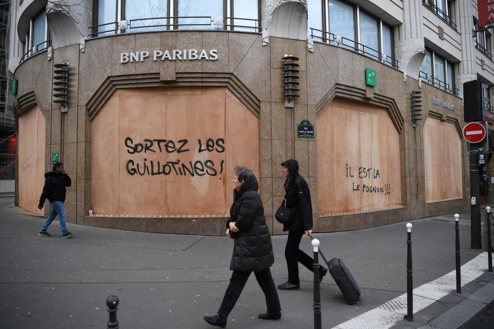 People walk past the protective wooden walls on a bank with a tag ,Get the guillotines out, and ,money is here, on Boulevard des Capucines in Paris, Dec. 9.