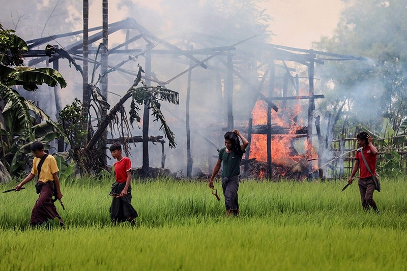 In this photograph taken on September 7, 2017, unidentified men carry knives and slingshots as they walk past a burning house in Gawdu Tharya village near Maungdaw in Rakhine state in Northern Myanmar (AFP Photo)